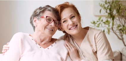 Smiling-nurse-in-beige-uniform-hugging-old-lady-drinking-tea-while-sitting-on-white-sofa-in-nursing-home